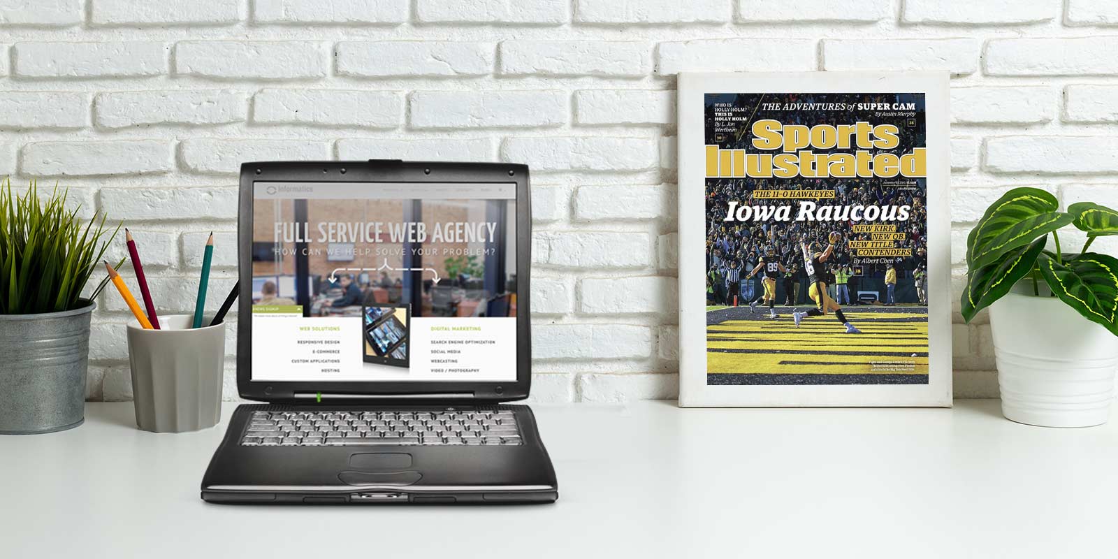 An old computer shows the Informatics website as it appeared in 2015 on a desk, while a poster for the Iowa Hakweyes football team appears in the background